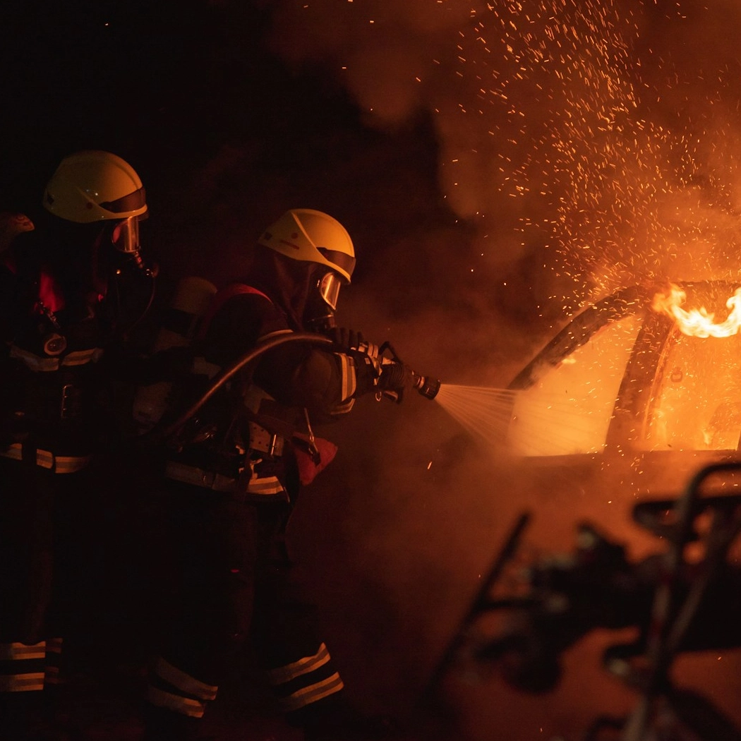 Two firefighters wearing breathing apparatus spraying water on a burning car at night