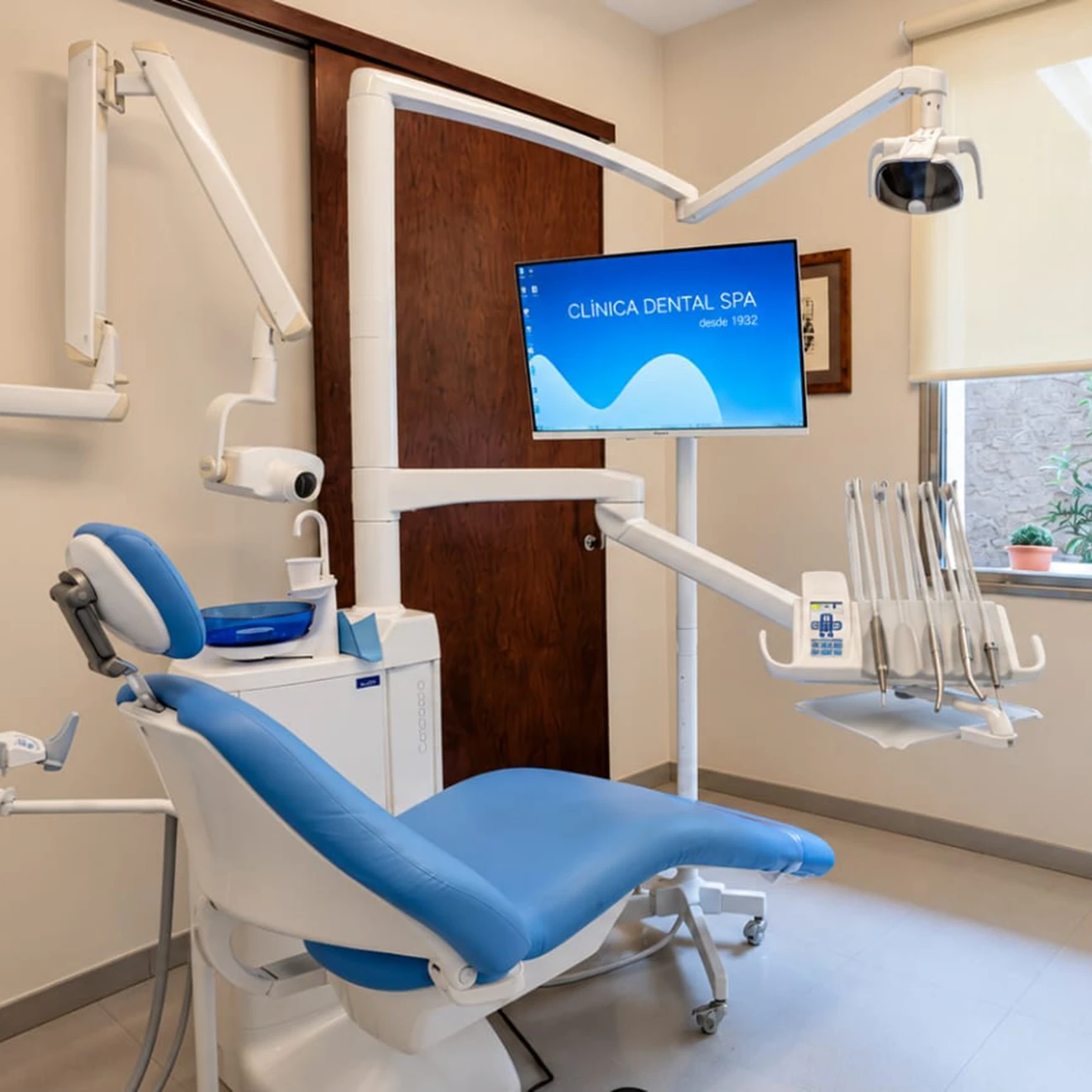 Clínica Dental SPA treatment room with a reclined blue dental chair, an articulated overhead light and a chairside monitor displaying the clinic's branding.